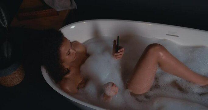 Close-up of young relaxed happy attractive African American female enjoying foamy bath and using smartphone.