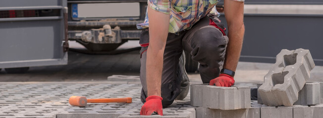 Mistrz brukarski układa ( brukuje) podjazd za pomocą kostki betonowej z otworami ( ażurowych bloczków betonowych). Widoczny bezwładnościowy młotek z tworzywa sztucznego - pomarańczowy kolor.