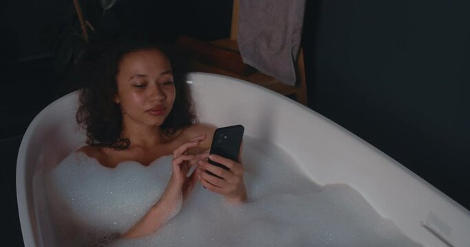 Enjoying vacation. Happy cheerful attractive young woman relaxing in elegant bath using smartphone.