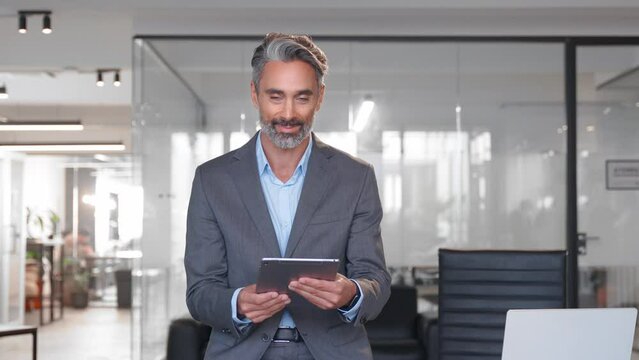 Happy Latin Hispanic bearded stylish mature adult professional business man, smiling Indian senior businessman CEO holding digital tablet using fintech tab application standing inside company office.
