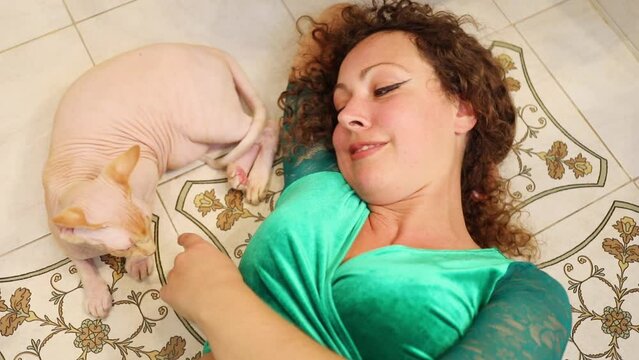 Angry sphinx cat and woman lie on floor in room, cat bites woman