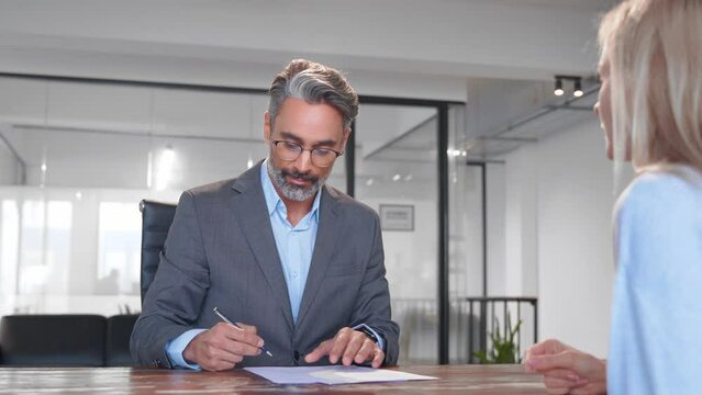 Mature Latin businessman, European businesswoman discussing project, sign contract, handshake sitting at table in office, slowmo. Business people work together. Agent man make consultation for client
