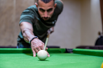 Young adult bearder male playing billiards with his friends focusing on white ball to hit it with...