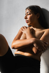 Fototapeta premium brunette latina woman covering her breasts with her hands folded while leaning against the wall with a sad look looking down.