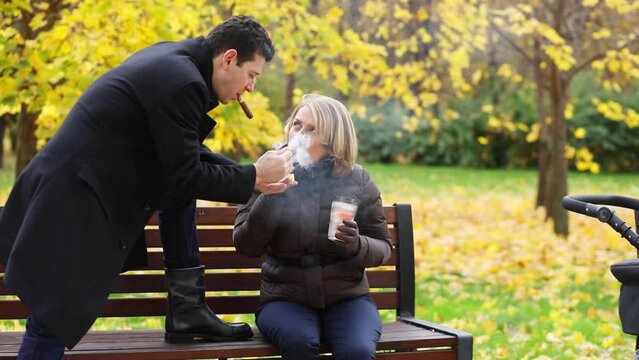 Handsome man with cigar lights cigar for woman on bench in autumn park