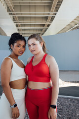 Portrait of two plus-size female athletes standing outdoors and looking at camera. Two young females in fitness attire standing together.
