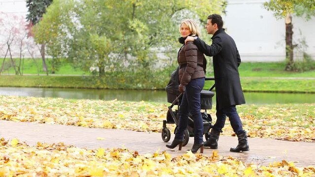 Happy man and pretty woman walk with baby carriage near river in fall park