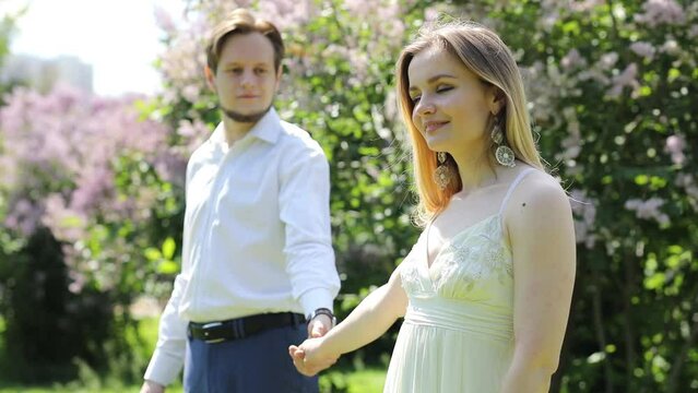 Happy girl calls man in park with lilac at sunny day, slow motion, focus on woman