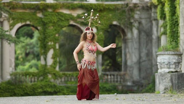 Pretty woman in red costume dances belly dance with chandelier on head in garden of sanatorium, 