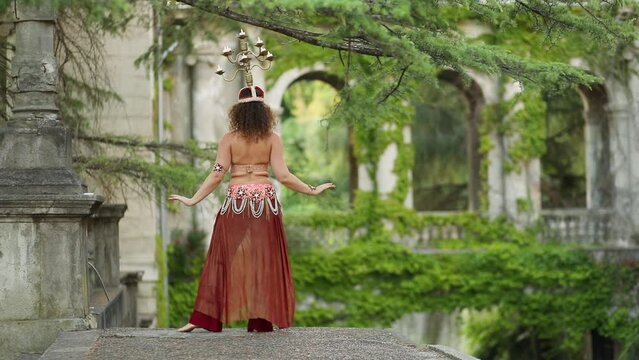 Pretty woman in red costume dances belly dance with chandelier on head in green garden of sanatorium, 