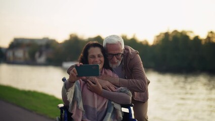Wife in wheelchair makes couple selfie. Senior happy family spending time in green city park taking photos on smartphone. Man hugging woman showing love care. Living with physical disability. - Powered by Adobe