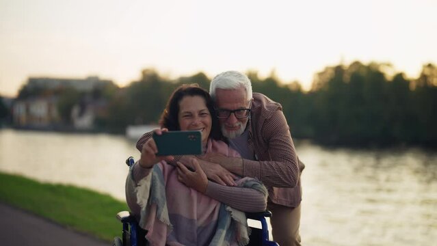 Wife in wheelchair makes couple selfie. Senior happy family spending time in green city park taking photos on smartphone. Man hugging woman showing love care. Living with physical disability.