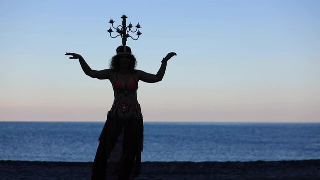 Woman dances belly dance with chandelier on head near blue sea at evening