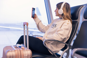 Relaxed attractive caucasian young woman using a smartphone to take a picture at the airport with suitcase next to her and headphones waiting for boarding