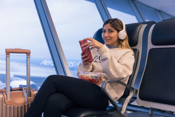 Relaxed attractive caucasian young woman using a smartphone and eating candies at the airport with suitcase next to her and headphones looking and smiling at the camera