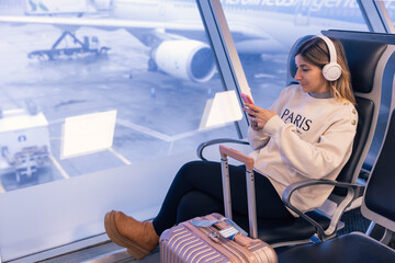 Relaxed attractive caucasian young woman using a smartphone at the airport with suitcase next to her and wearing headphones