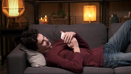Young happy Caucasian man is texting on phone, participating in an online date, smiling while lying on the sofa in the living room. Dating app, lovers conversation, online messaging.