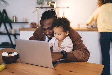 Focused black father with cute little daughter typing laptop while sitting at dining table