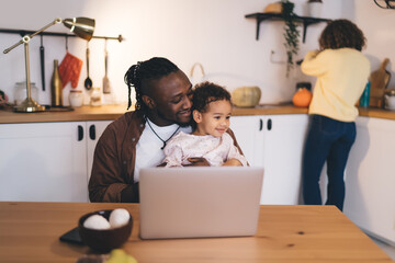 Smiling ethnic daughter carried by father while sitting at dining table and using laptop in kitchen