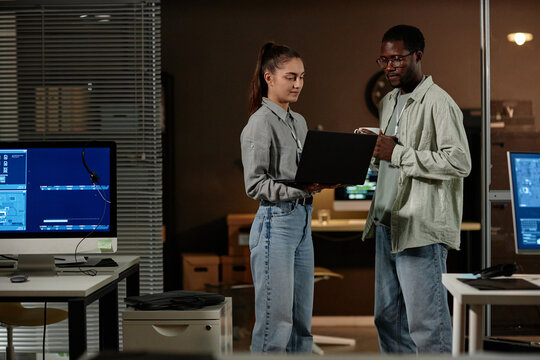 Portrait of two young people using laptop while working in IT office at night, copy space