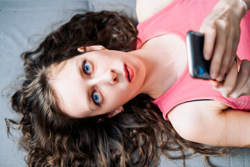 Young woman with long curly hair lies on the sofa and scrolls social networks on her phone in a...