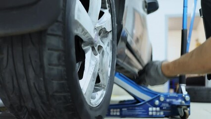 Worker tightens wheel of car which stand on lifting jack in service garage. Slow motion