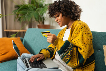 African American woman using laptop shopping online paying with gold credit card. Girl sitting at...