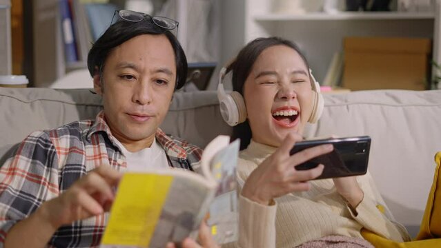 asian female woman having fun playing online game smartphone and husband reading book sit near together in living room at home.concept of happiness couple,good time,relative family,love couple
