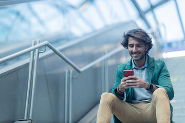 View of young man sitting down using a smartphone at indoor space with a blurred view landscape in the background. High quality photo. Texting on the phone. 