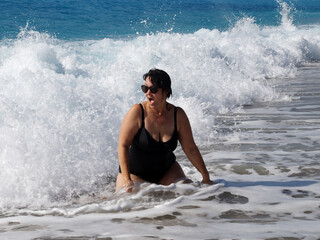 curvy woman in a black swimsuit and sunglasses sits in the sea under the lapping waves