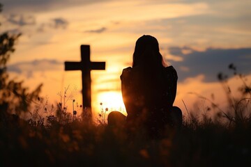 Silhouette of a woman sitting on the grass and praying in front of a cross at sunset. - obrazy, fototapety, plakaty