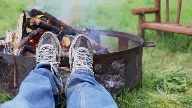 Mans legs in jeans and sneakers are near burning campfire on grass near wooden bench. Slow motion