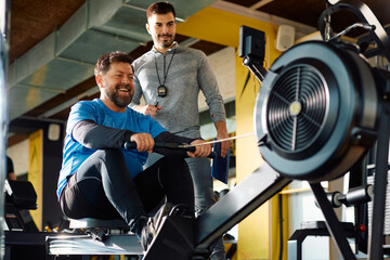 Happy mature man working out on rowing machine while having exercise class with coach in gym.