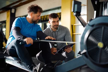 Fototapeten Young coach and mature athlete going through exercise plan during workout on rowing machine in gym. © Drazen