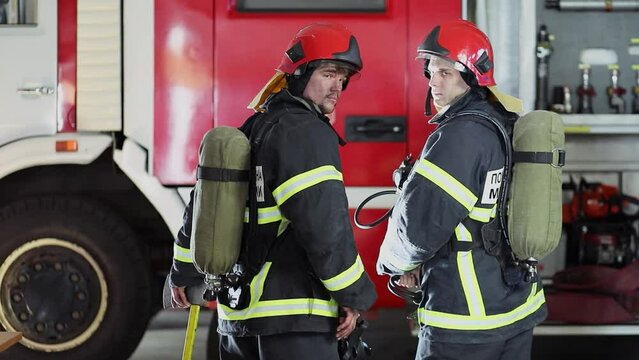 Two firefighters turn around in protective coats with oxygen cylinders near fire truck