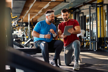 Fitness instructor using touchpad while showing exercise plan to middle aged athlete in gym.