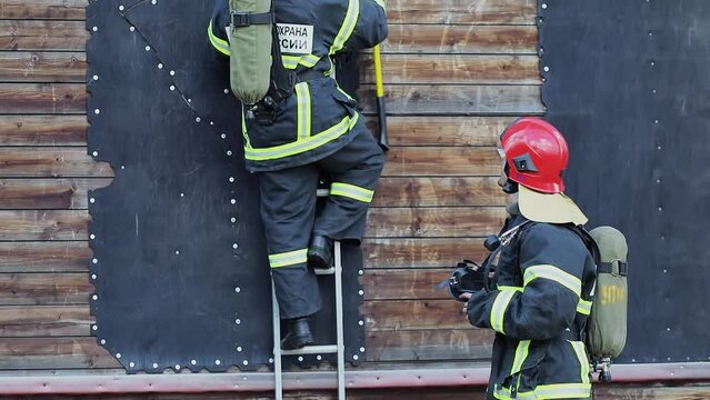 Fireman in protective suits with russian inscription Fire Guard of Russia Emercom, climbs by ladder on training wall and his partner stands near. Slow motion