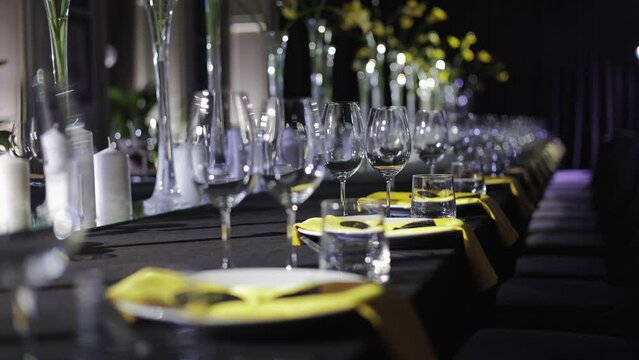 Served decorated black table with sparkling empty wineglasses, porcelain platters and bright details in form of yellow flowers and napkins. Side view of violet empty armchairs standing by dining area.