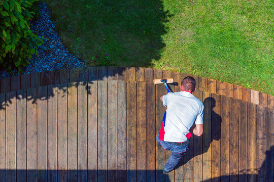 Terrace cleaning. Worker renovating a gray terrace using a hard scrub brush and green gel for renovating faded, gray boards - an alternative to washing with a high-pressure washer