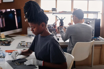 Black woman with braids repairing cellphone at workshop desk, her coworker sitting on background