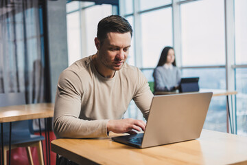 Fototapeta premium A young successful man works in a bright office sitting at a table with a laptop. Freelance work. Remote work in a cafe.