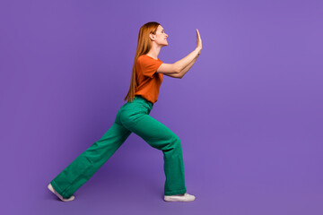 Full length size photo of a hardworking woman in green pants and orange t shirt pushing object isolated on violet color background