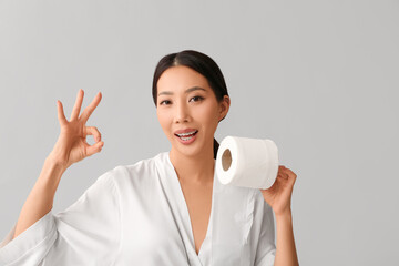 Young Asian woman in gown with toilet paper showing OK on light background