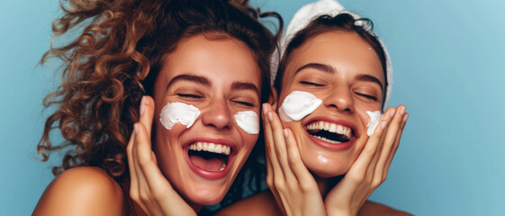 Two joyful women with face cream, sharing a moment of beauty routine fun against a blue background