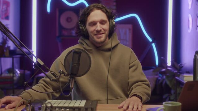 UGC of Caucasian bearded man looking at camera and speaking into microphone sitting at table with professional musical equipment and recording blogger content at neon lit home studio at night