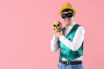 Happy young man in costume and carnival mask holding painted human skull on pink background