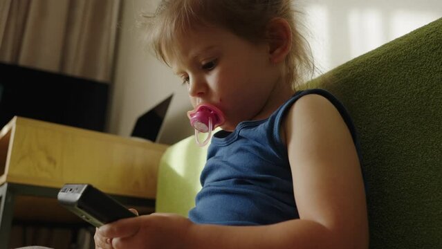 Little kid girl sucking a pacifier while sitting and playing game portable device at home. Tech kids addiction.