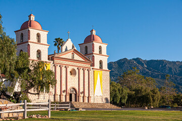 Fototapeta na wymiar Santa Barbara, CA, USA - April 20, 2009: Old Mission church front facade in sun under blue sky. Green trees and mountains in back. The Santa Barbara Mission was established on the Feast of Saint Barb