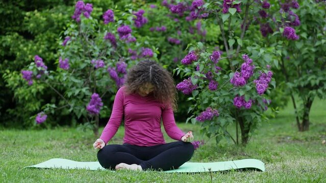 Woman sits on rug and rotates head near bush of blooming lilac. Slow motion