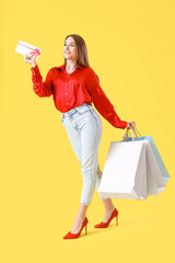 Happy young woman with gift card and shopping bags on yellow background